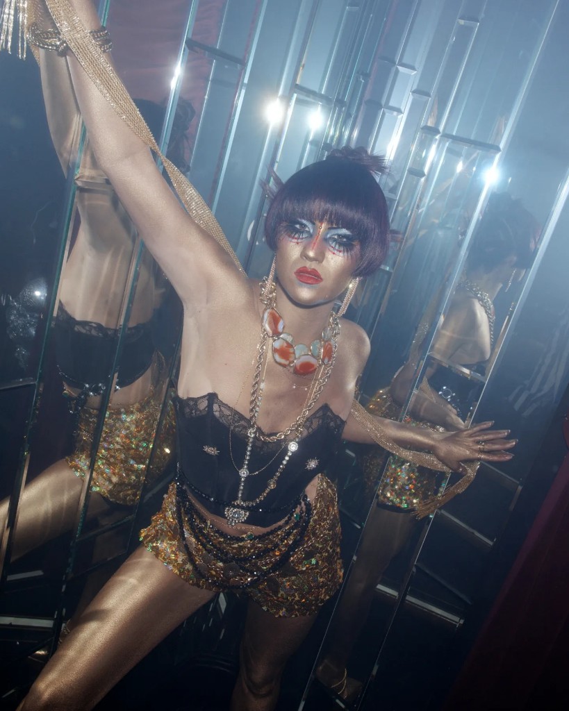 Gold body and graphic eye makeup in a mirrored room